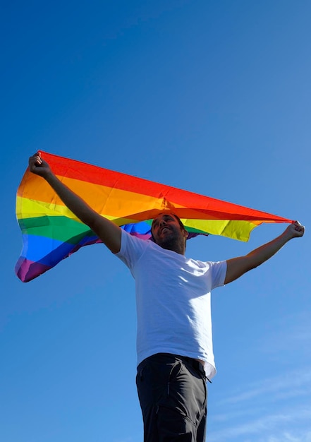 Erwachsener Mann im weißen T-Shirt winkt Regenbogenfahne mit blauem Himmel.