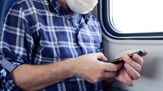 Erwachsener Mann in einem Pendlerzug mit Kopfhörern und einer medizinischen Maske mit einem Telefon, das Musik hört