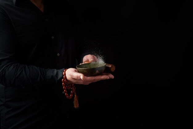 Erwachsener Mann in einem schwarzen Hemd dreht einen hölzernen Stock um eine kupferne tibetanische Schüssel Wasser