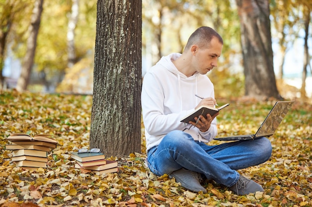 Erwachsener Mann in einem weißen Kapuzenpulli studiert im Park auf einem Laptop, schreibt in ein Notizbuch, liest Bücher und Lehrbücher