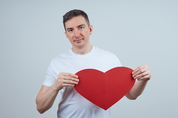 Erwachsener Mann in einem weißen T-Shirt, mit einem Papierherz in seiner Hand, auf einem grauen Hintergrund. Fröhlichen Valentinstag. Weltherztag.