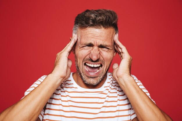 Erwachsener Mann in gestreiftem T-Shirt schreit und reibt sich die Schläfen wegen Kopfschmerzen isoliert auf Rot