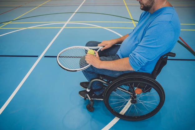 Erwachsener Mann mit einer körperlichen Behinderung, der den Rollstuhl benutzt, um Tennis auf einer Tennishalle zu spielen