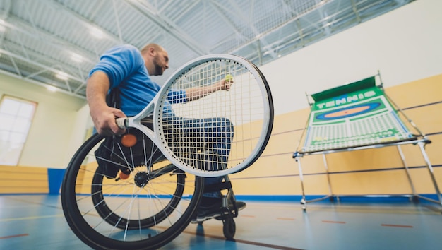 Erwachsener Mann mit einer körperlichen Behinderung, der den Rollstuhl benutzt, um Tennis auf einer Tennishalle zu spielen