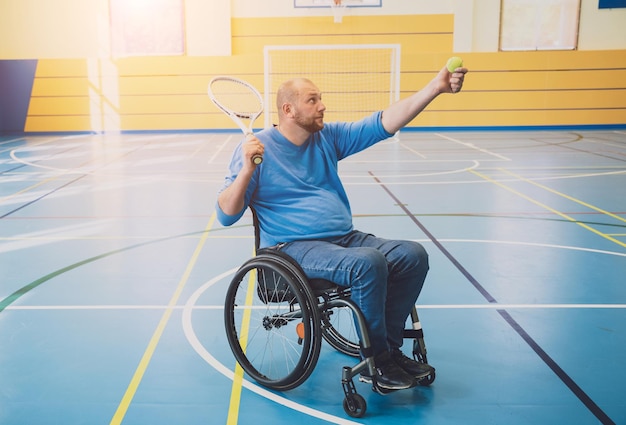 Erwachsener Mann mit einer körperlichen Behinderung, der den Rollstuhl benutzt, um Tennis auf einer Tennishalle zu spielen