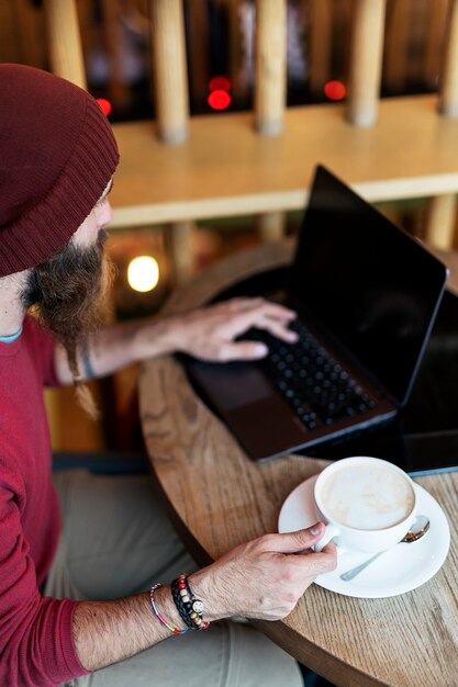 Erwachsener Mann mit geflochtenem Bart, der im Café sitzt und eine Tasse Kaffee trinkt, während er auf dem Laptop tippt