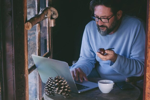 Erwachsener Mann sitzt zu Hause, arbeitet an einem Laptop und raucht eine Pfeife Alternativer Single-Lebensstil Männer in gemütlichen Hütten, hölzerne Chalet, Online-Telearbeit