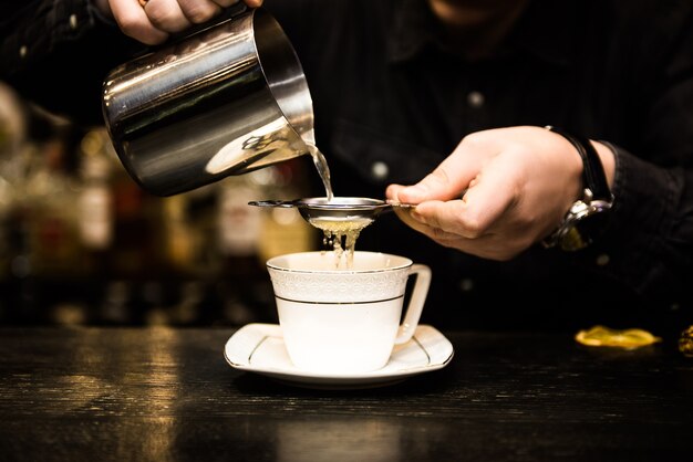 Erwärmender alkoholischer Cocktail in einer weißen Tasse auf einem dunklen Tisch, die Bar mit Zimtstange und Orangenorange. Der Kochvorgang