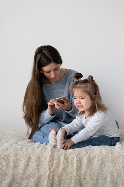 Es difícil trabajar de forma remota en casa. Hermosa joven madre se sienta en casa en un sofá. Teléfono móvil en las manos. Pequeña hija llorosa cerca de la madre. El niño esta sufriendo