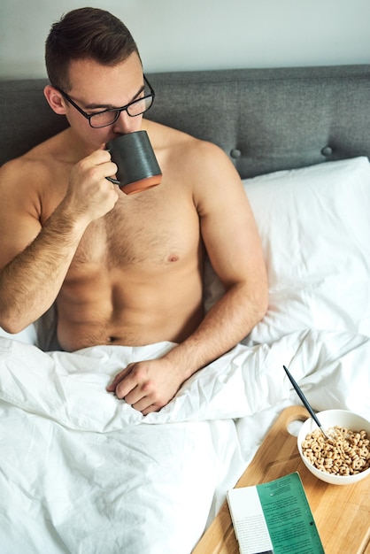 Esta es una excelente manera de comenzar la mañana Captura recortada de un apuesto joven sin camisa tomando café y desayunando en la cama en casa