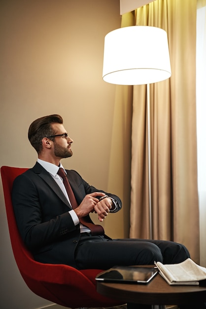 Es hora de descansar. Hombre de negocios esperando a alguien en la silla roja en la habitación del hotel. Mira su reloj de pulsera