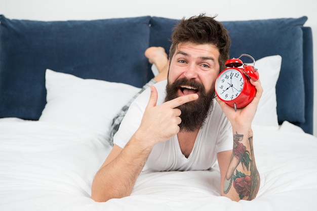 Es hora de despertarse Por qué debería levantarse temprano todas las mañanas Beneficios para la salud de levantarse temprano Despertarse temprano da más tiempo para prepararse y ser oportuno Hombre barbudo hipster acostado en la cama con despertador