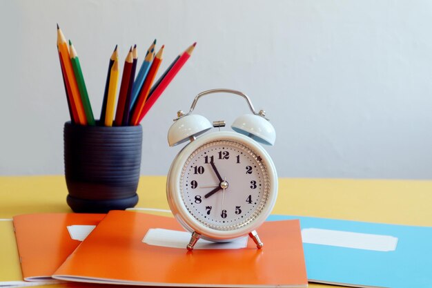 Es hora de ir a la escuela, un reloj de alarma blanco se encuentra en cuadernos multicolores con el fondo de lápices en un espacio de vista lateral de fondo gris de taza para texto