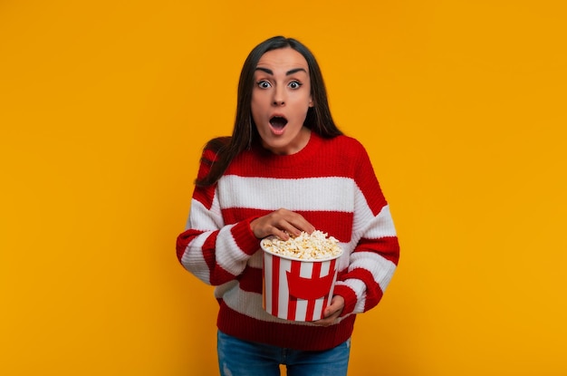 ¡Es imposible! Primer plano retrato de joven hermosa mujer morena en suéter mientras sostiene un gran cubo de palomitas de maíz para ver películas en el cine y sorprendida mirando a la cámara