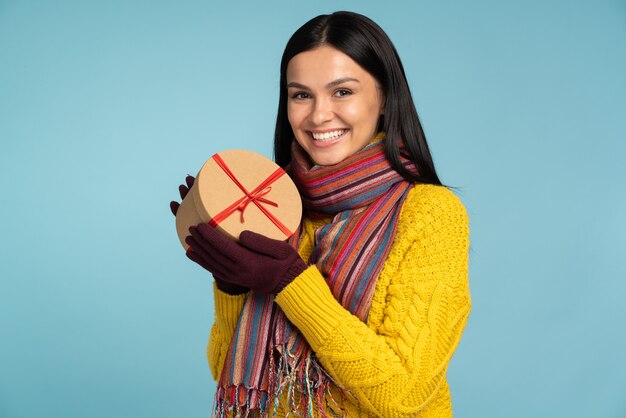 Es ist für dich. Lächelnde wunderschöne Frau in stilvollem gelbem Pullover mit Schrankgeschenkbox, während sie vor der Kamera steht und auf blauem Wandhintergrund posiert. Konzept der Winterferien