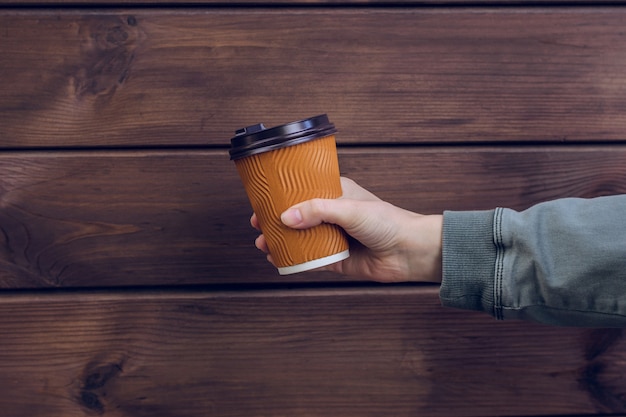 Es ist Kaffeezeit! Frauenhand, die Pappbecher des frischen Kaffees gegen dunklen hölzernen Hintergrund hält