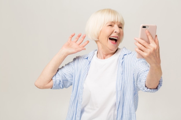 Es ist Selfie-Zeit! Bild der fröhlichen reifen alten Frau, die lokal über weißer Hintergrundwand steht, die durch Handy spricht.