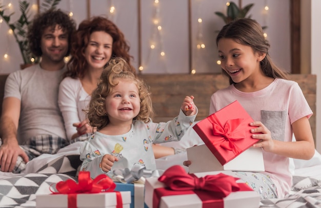 Es ist Weihnachtszeit! Glückliche Familie, die im Bett sitzt, Eltern lächeln, während ihre Töchter die Geschenke öffnen