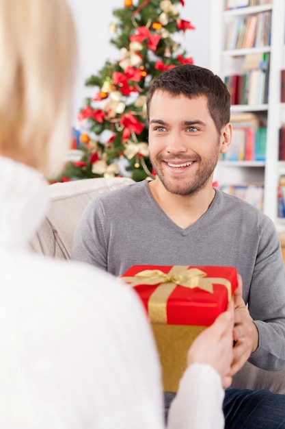 ¿Es para mi? Alegre joven sentado en el sofá y recibir una caja de regalo de su novia sentada cerca de él.