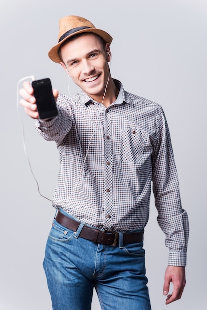 Foto ¡esta es mi canción favorita! apuesto joven en fedora mostrando su reproductor de mp3 y sonriendo mientras está de pie contra el fondo gris