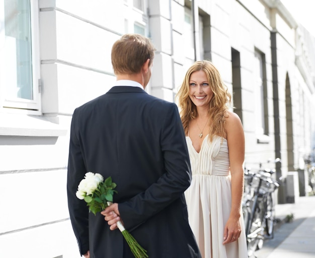 Es tan bromista Una hermosa mujer le sonríe a su novio sosteniendo un ramo de rosas blancas detrás de su espalda