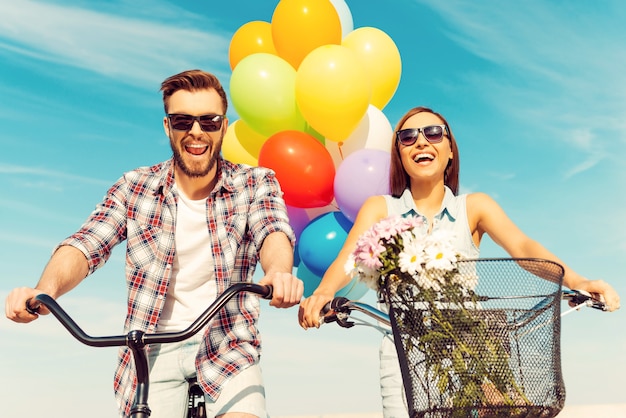 ¡Esto es tan divertido! Vista de ángulo bajo de la alegre pareja joven sonriendo y montando en bicicleta con globos de colores en el fondo