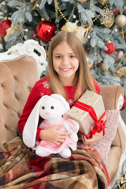 Es tan tierno y tierno. Niño pequeño feliz sonriendo con regalos. Niña con lindo conejito en el árbol de Navidad. Niña pequeña mantenga el juguete del conejo. Juego de niño pequeño con peluche. Mejor juguete navideño.