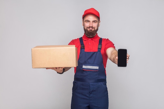 ¡Esto es para ti! Sonriente joven logístico con barba en uniforme azul y camiseta roja de pie, sosteniendo una caja de cartón y mostrando la pantalla del teléfono inteligente sobre fondo gris. Interior, tiro del estudio aislado.