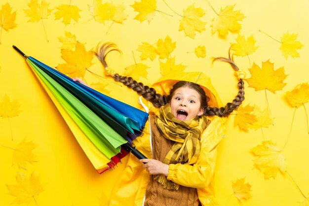 Es tiempo de otoño para una niña pequeña con coletas y una bufanda El niño está acostado sobre hojas de arce amarillas con un paraguas multicolor en un suéter impermeable y sonriendo ampliamente