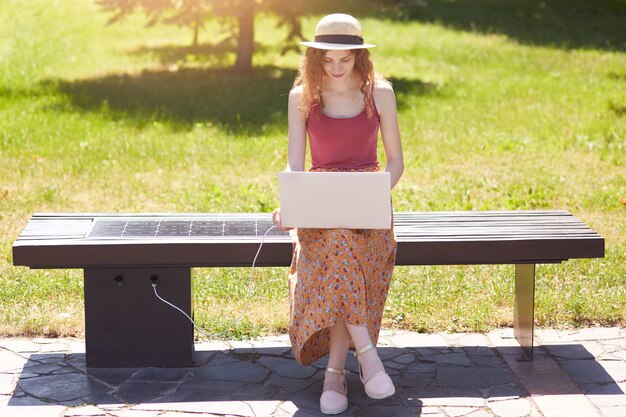 Esbelta chica atractiva que busca información en la red, carga portátil con ayuda de un panel solar construido en un banco de madera, aprovecha las ventajas de las nuevas tecnologías, está de buen humor. Concepto ecológico.