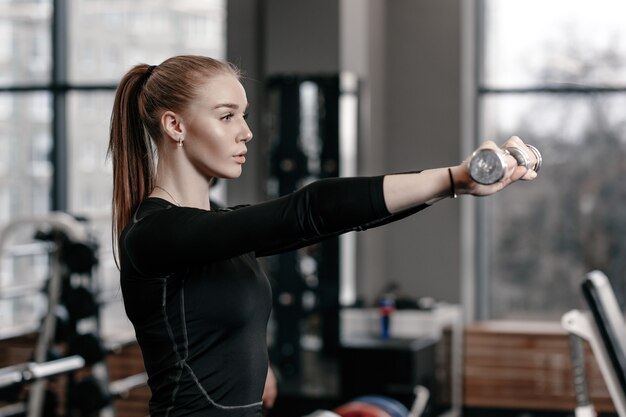 Esbelta joven vestida con ropa deportiva negra está haciendo ejercicios con pesas en el moderno gimnasio con una gran cantidad de equipamiento deportivo.