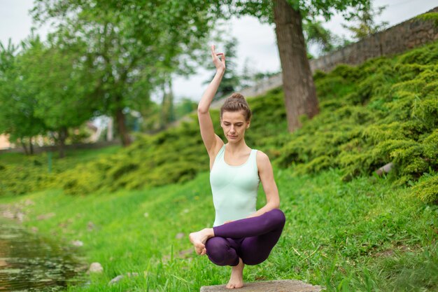 Esbelta joven yogui morena realiza ejercicio complejo de yoga en la hierba verde.