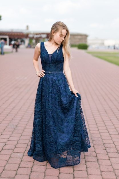 Esbelta loira tocando a bainha do elegante vestido azul escuro do chão na calçada.