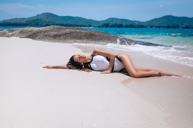 Una esbelta modelo en traje de baño blanco miente y toma el sol en la playa