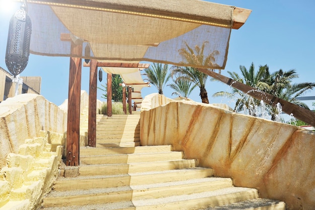 Escada ao ar livre com uma barraca de sol ao fundo há palmeiras e jatos de água uma lâmpada típica árabe está pendurada