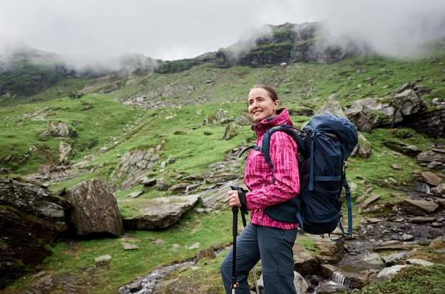 Escalador femenino con bastones y mochila posando en la ladera rocosa verde hierba