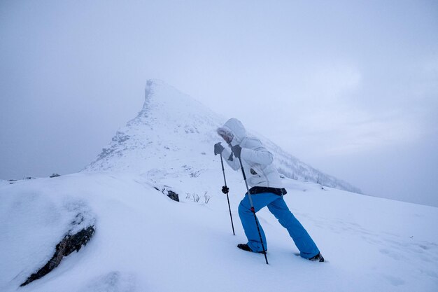 Escalador de hombre con bastones de trekking de pie en el pico de la montaña nevada en sombrío en noruega
