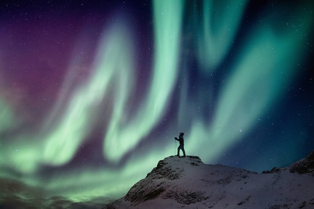 Escalador del hombre de pie en el pico nevado con aurora boreal y fondo estrellado