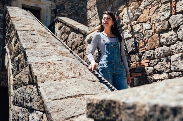 Escalera decadente en un antiguo castillo medieval histórico abandonado. joven turista asiática en monos caminando por las escaleras de piedra visitando el antiguo palacio al aire libre en un día soleado sosteniendo la cuerda.