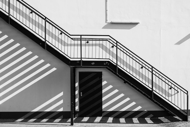 Una escalera exterior proyecta sombras sobre la pared y el suelo pavimentado, en blanco y negro.