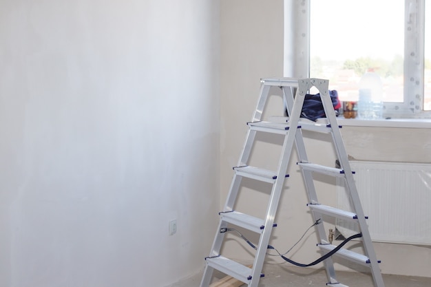 Escalera plegable de aluminio en una habitación con pared y ventana enlucidas en blanco. Reforma de vivienda en casa nueva. Copie el espacio.