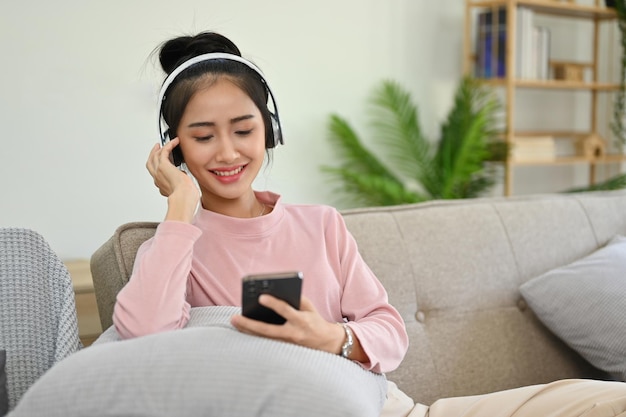 Escalofriante mujer asiática relajándose en la sala de estar con auriculares escuchando música o podcast