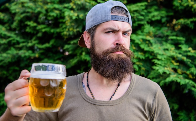 Escapando do calor do verão. Hipster brutal bebendo cerveja refrescante num dia de verão. Homem barbudo segurando a caneca de cerveja na natureza de verão. Bebedor aproveitando o horário de verão.