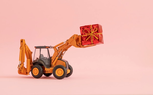 Escavadeira de brinquedo carrega uma caixa de presente amarrada com um laço. fundo rosa copie o espaço,