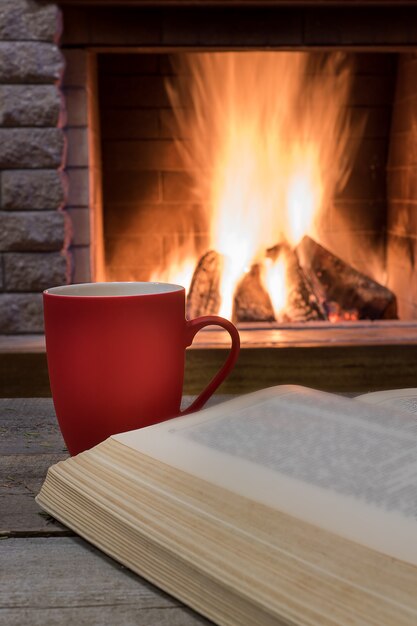 Escena acogedora antes de la chimenea con una taza roja de té, y libro abierto.