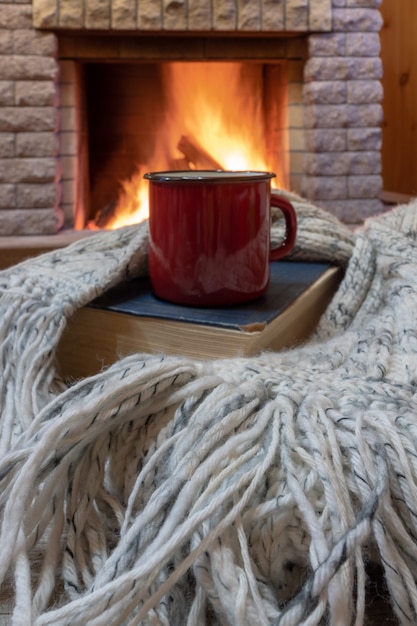 Escena acogedora antes de la chimenea con taza roja con té, un libro, bufanda de lana.