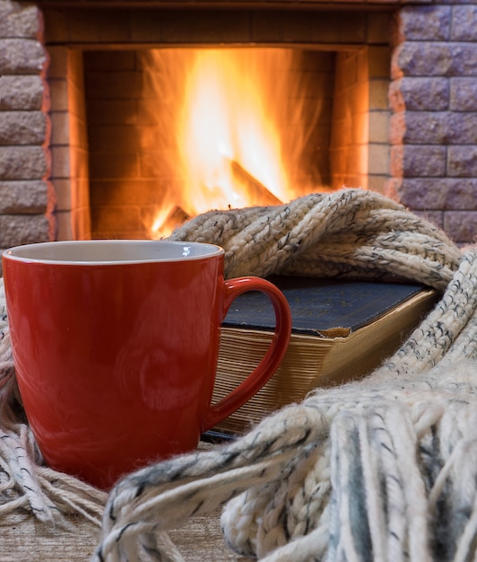 Escena acogedora antes de la chimenea con una taza roja de té, un libro, bufanda de lana.