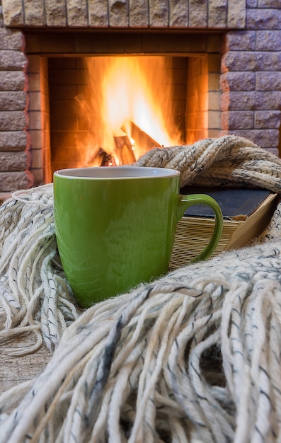 Escena acogedora antes de la chimenea con taza verde con té, un libro, bufanda de lana.