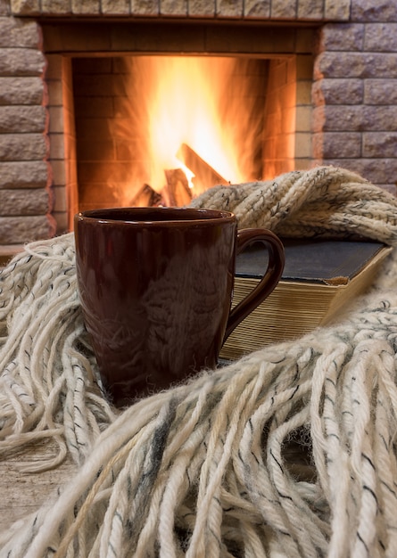 Escena acogedora ante chimenea con taza marrón con té, libro, bufanda de lana.