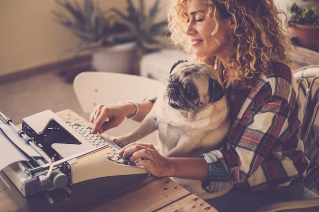 Escena divertida con hermosa dama rubia rizada hipster trabajando y escribiendo en una vieja máquina de escribir escribiendo un blog o libro mientras su mejor amiga ama el viejo perro pug escribe lo mismo y bromea con su dueño